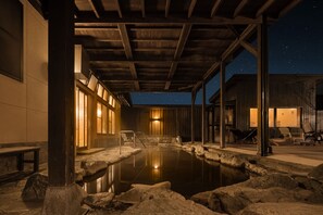 A night-time hot spring area with a wooden roof, stone seating, and a view of the night sky.