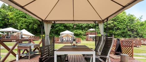 An outdoor wooden deck with a table and chairs under a canopy, surrounded by wooden fences and tents.