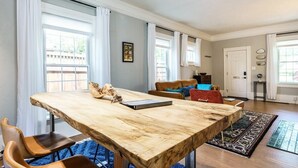 A spacious living room with a large wooden dining table, comfortable seating, and large windows with curtains.