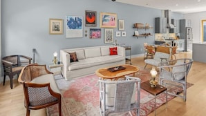 Ein modernes Wohnzimmer mit einem weißen Sofa, Korbstühlen, einem Couchtisch und einer Küchenzeile mit Schränken und Geräten.