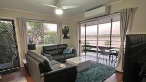 Un salon moderne avec un canapé, une table basse et un téléviseur, de grandes fenêtres offrant une vue sur un plan d’eau, et un ventilateur de plafond.