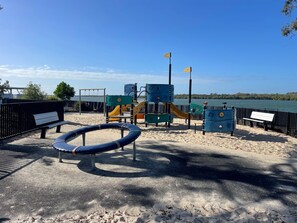 Children can play on the swings, slides, and climbing structures in this fenced outdoor play area overlooking a serene body of water.