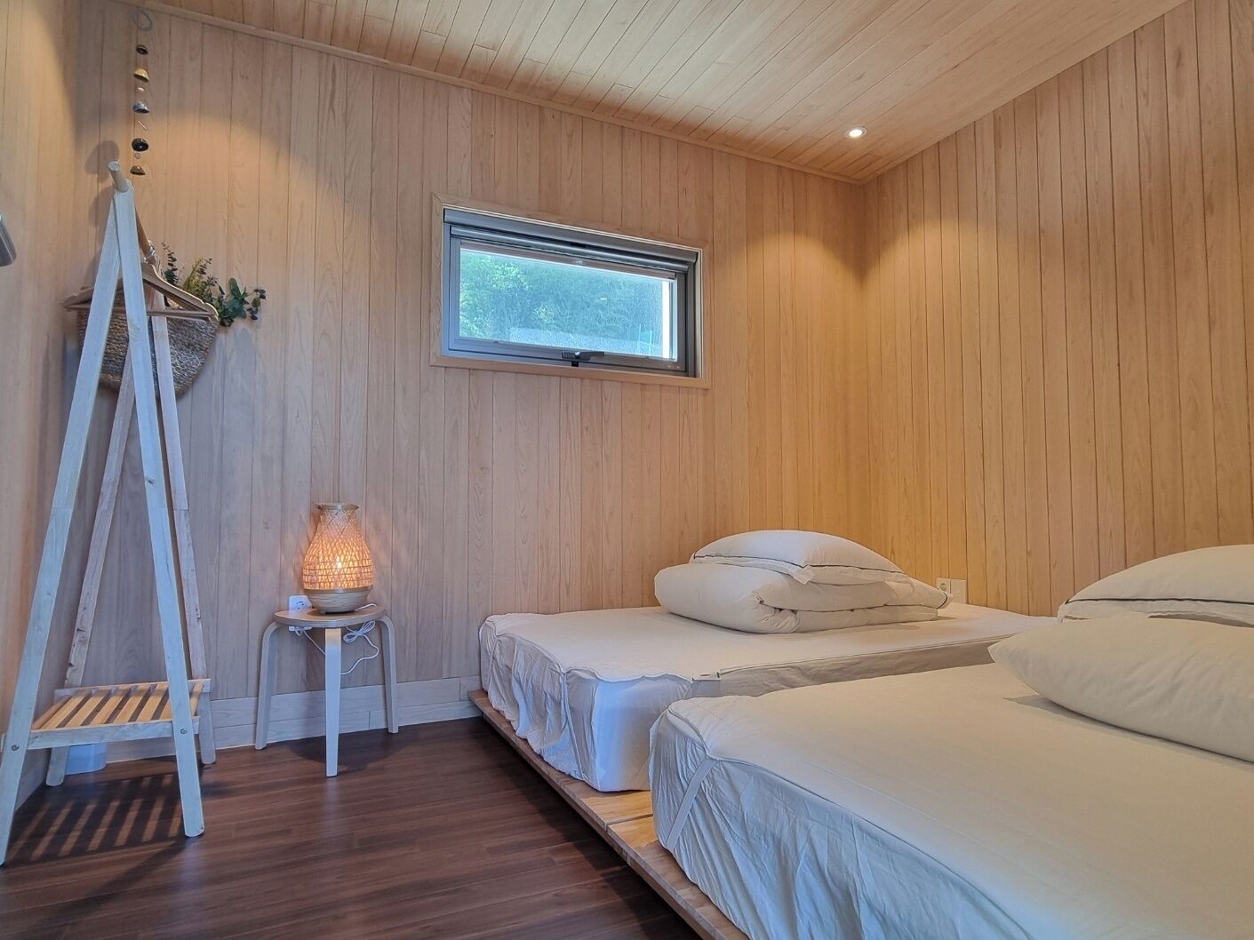 A bedroom with wooden walls, a window, a ladder, and a bedside table with a lamp.