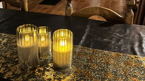 A cozy dining area with a table set for two, featuring a black tablecloth and golden glittery placemat. There are three lit candles on the table. In the background, there is a wooden dining table with chairs, a fireplace with a log fire, and a wall with musical notes and a clock. - House overlooking the Cadí-Moixeró massif, traditional from the 70s with magical (Lles de Cerdanya)