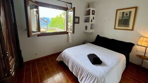 A bedroom with a bed, a window with a view, a bedside table, and a wall-mounted shelf with decorative items.