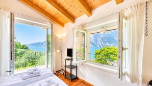 A bedroom with a bed, a TV on a stand, and a balcony with a view of mountains and the sea. - Villa Il Paradiso Di Gabri: Lake-view Villa With Garden (Nago-Torbole)