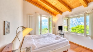 A bedroom with a wooden ceiling, a bed, a desk with a lamp, and a view of mountains through large windows. - Villa Il Paradiso Di Gabri: Lake-view Villa With Garden (Nago-Torbole)
