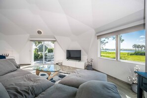 Ein modernes Wohnzimmer mit einem großen Sofa, einem Glastisch und einem Flachbildfernseher. Ein großes Fenster bietet Blick auf eine Rasenfläche und ein Gewässer.