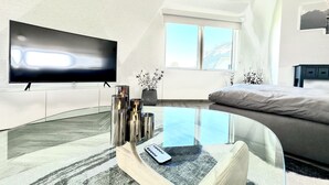 Ein modernes Wohnzimmer mit Flachbildfernseher, Glastisch, Sofa und Bett.