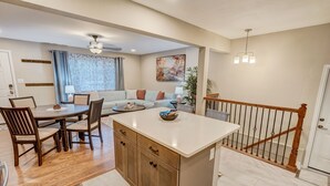 A modern kitchen with a central island, wooden dining table, and chairs. There is a staircase with a wooden railing leading to another level.