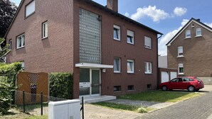 Un edificio de ladrillo de dos pisos con un garaje, un coche rojo estacionado afuera y un pequeño jardín.