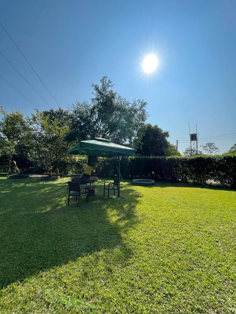 A grassy outdoor area with a table and chairs under a green canopy, surrounded by trees and a clear sky.