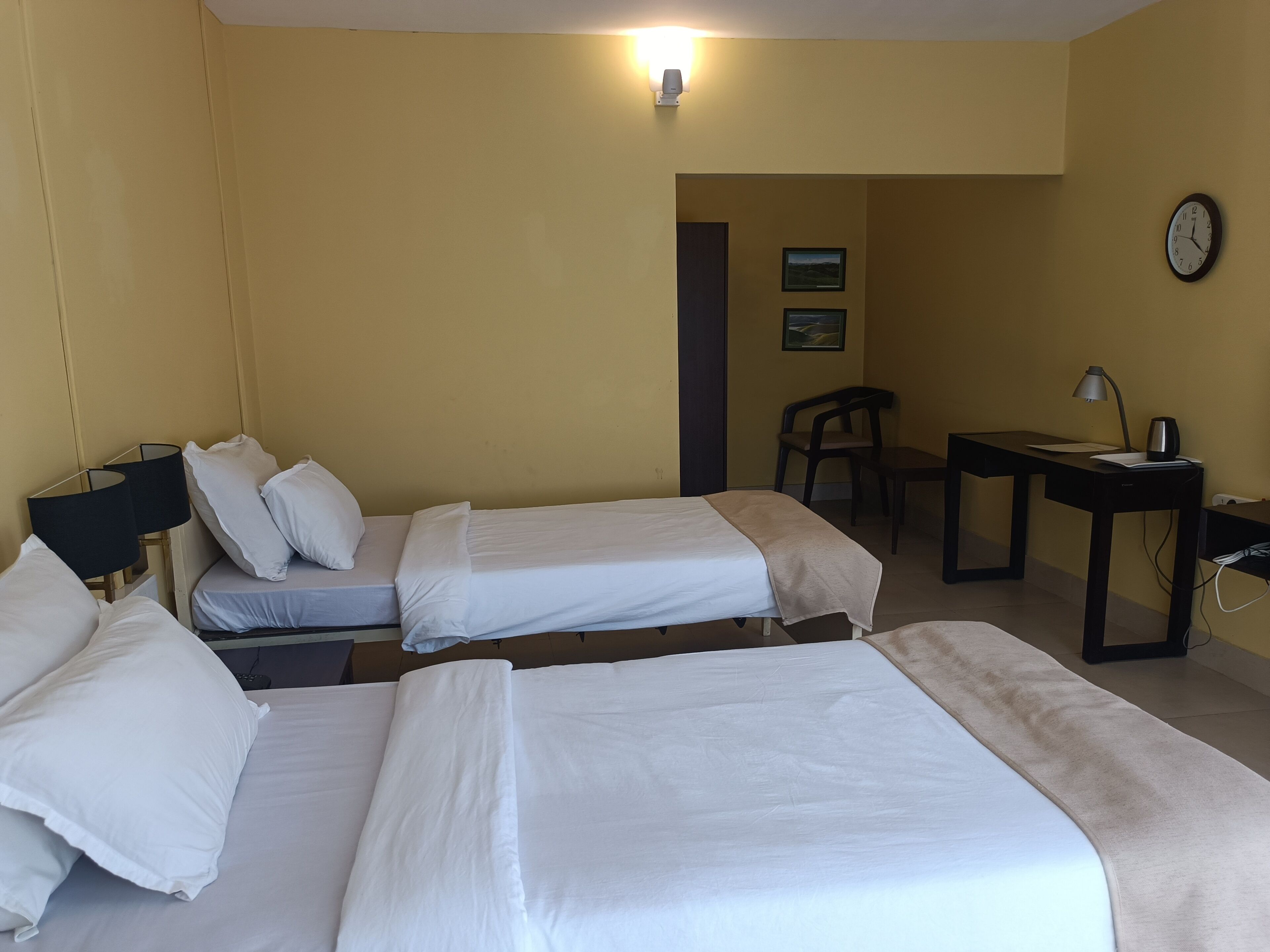 Une chambre d’hôtel avec deux lits, un bureau, une chaise, une horloge et une lampe fixée au mur.