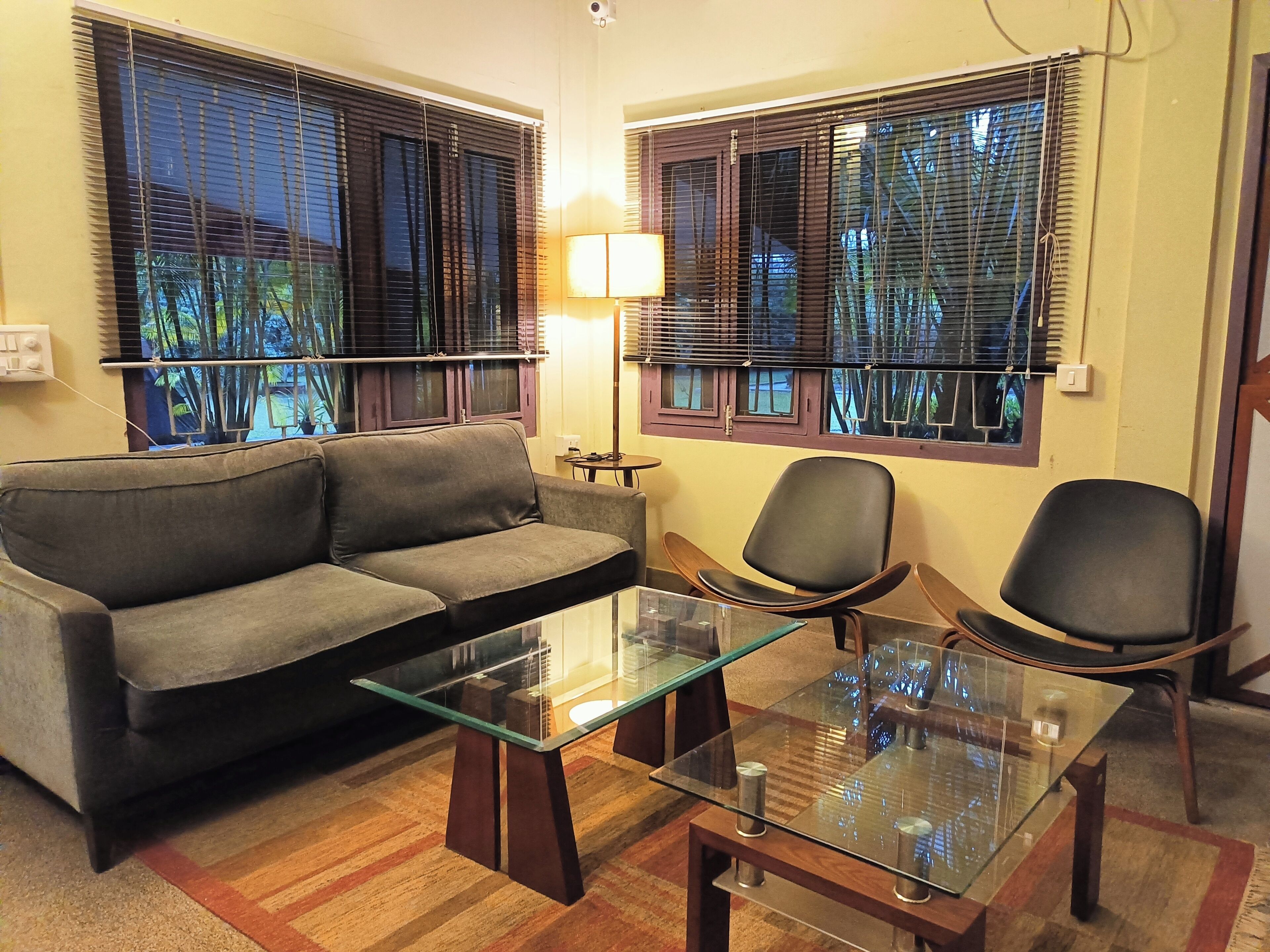 A room with a sofa, two chairs, a glass-top table, and windows with blinds.