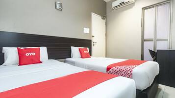 A hotel room with two beds, each with white bedding and red accents, a desk, and an air conditioning unit on the wall.