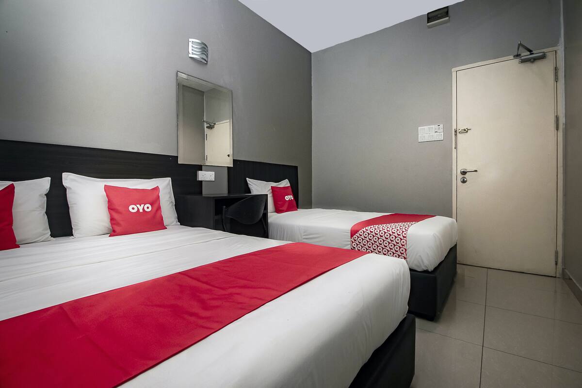 A hotel room with two beds, each with white bedding and red accents, a desk with a chair, a mirror, and a door.