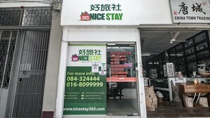 Entrada de hotel com placa indicando Nice Stay, oferecendo serviços como recepção 24 horas, TV com Astro e canais de filmes, e armazenamento de bagagem.