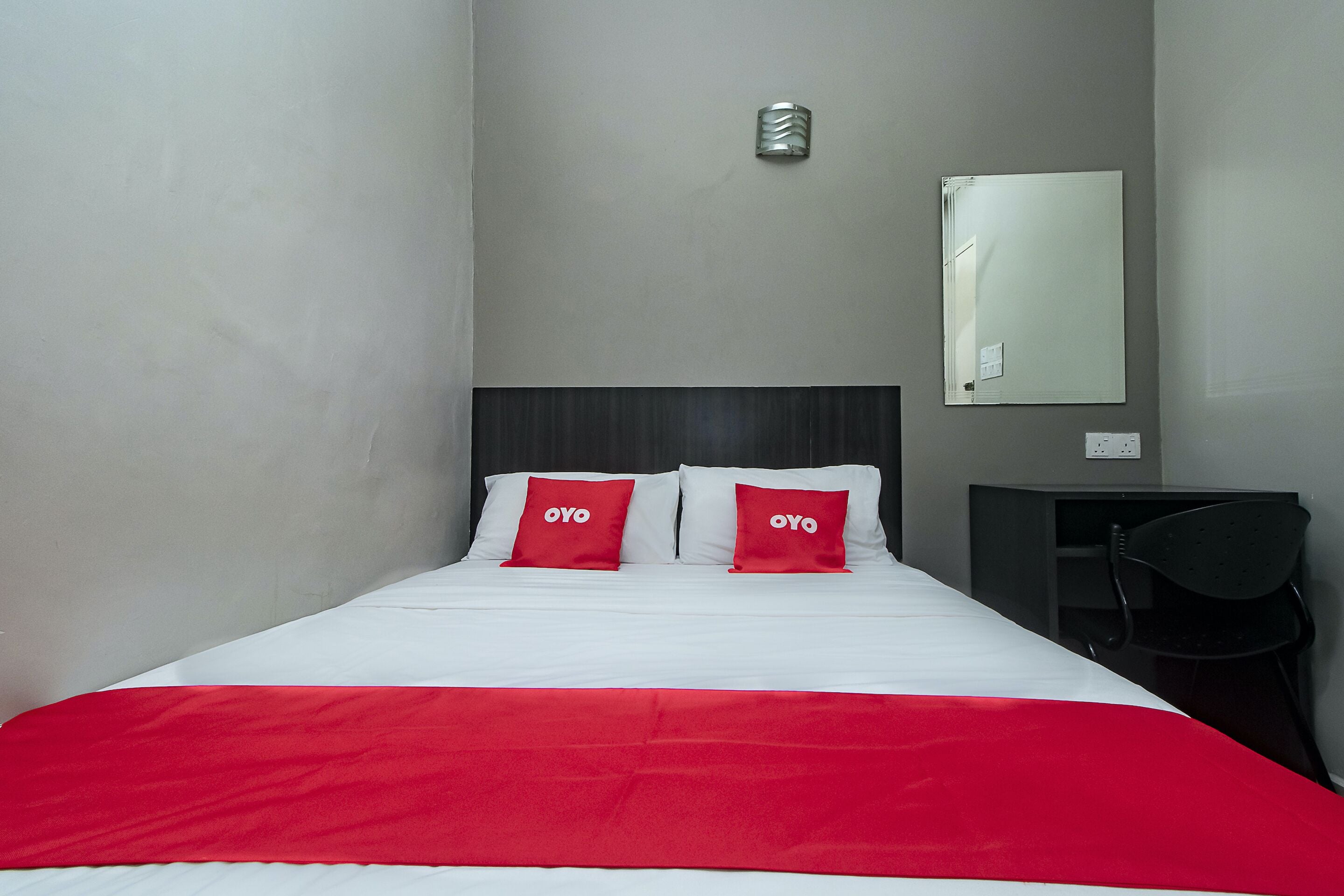 Une chambre d’hôtel avec un lit, deux oreillers rouges, une couverture rouge, une tête de lit sombre, un petit bureau avec une chaise et un miroir au mur.
