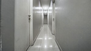 Um corredor comprido e estreito, com paredes brancas e chão de azulejos brilhantes, que leva até uma porta no final.