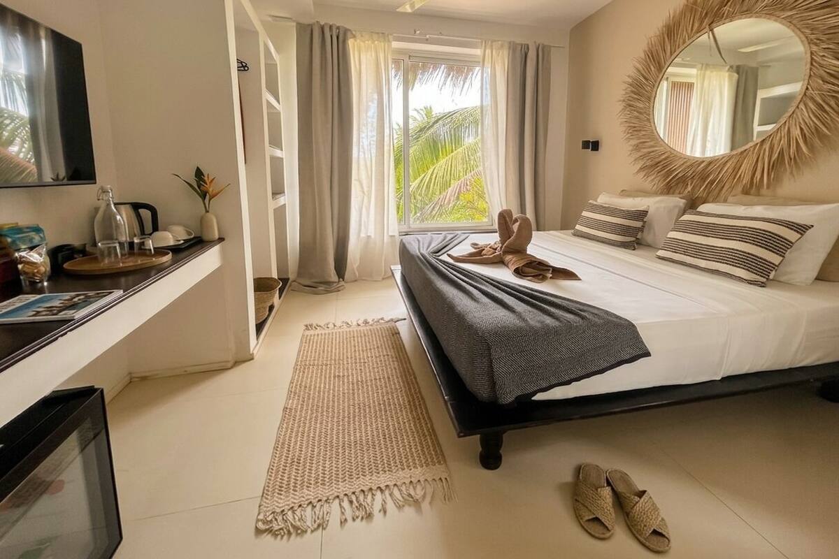 A bedroom with a bed, a TV, a mirror, a window with curtains, and a view of greenery outside.
