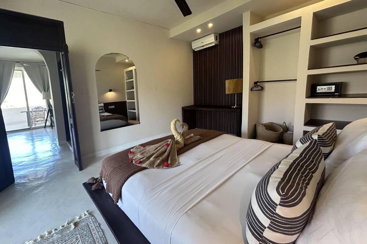 A modern hotel room with a large bed, a built-in wardrobe, a seating area with a view, and a balcony.