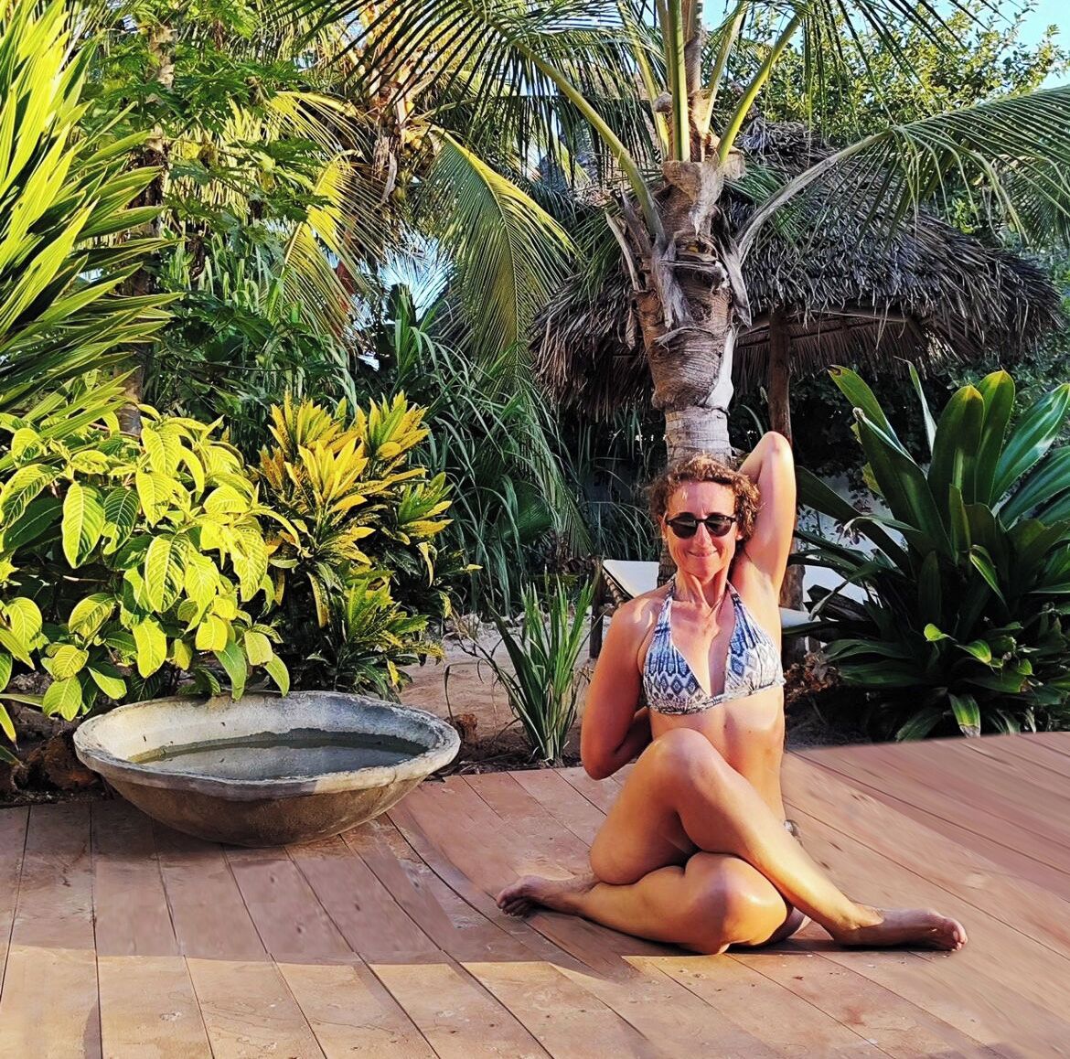 A person sitting on a wooden deck in a tropical setting with palm trees and various plants.