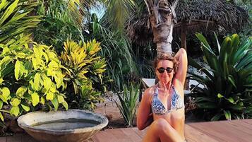 A person sitting on a wooden deck in a tropical setting with palm trees and various plants.