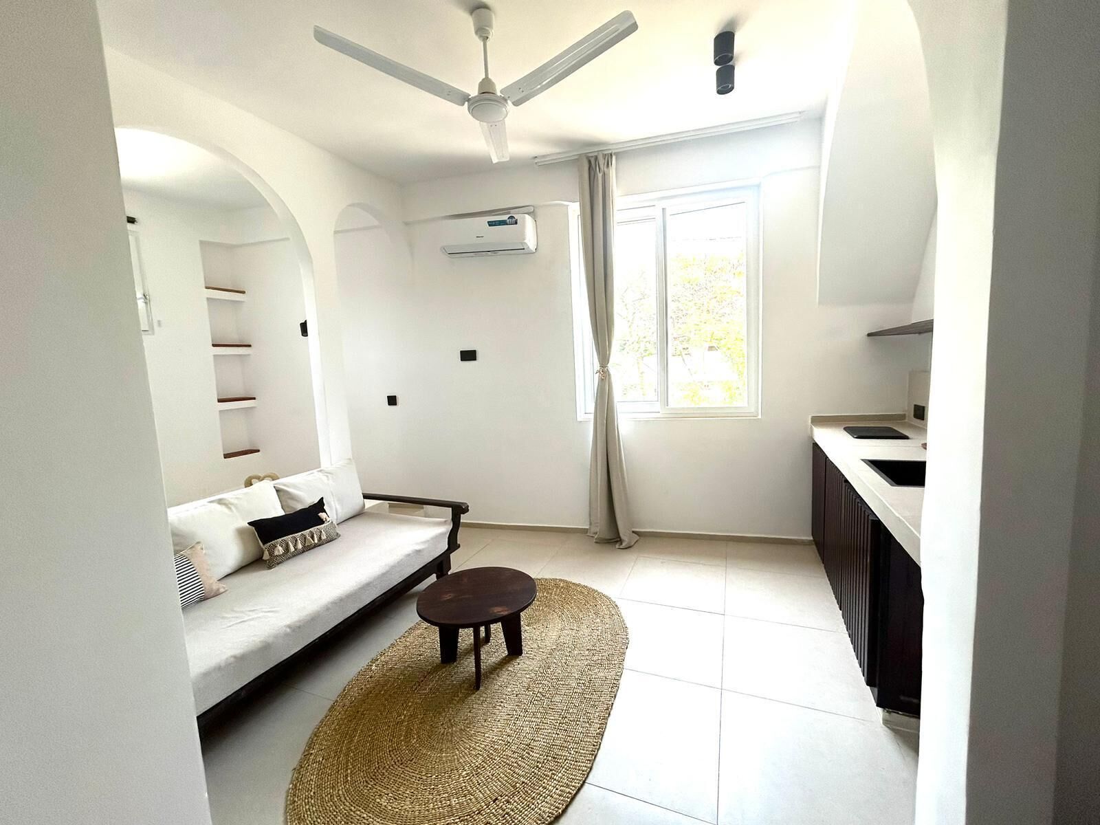 Un espacio habitable pequeño con sofá, una mesita redonda, ventilador de techo, aire acondicionado y una ventana con cortinas.