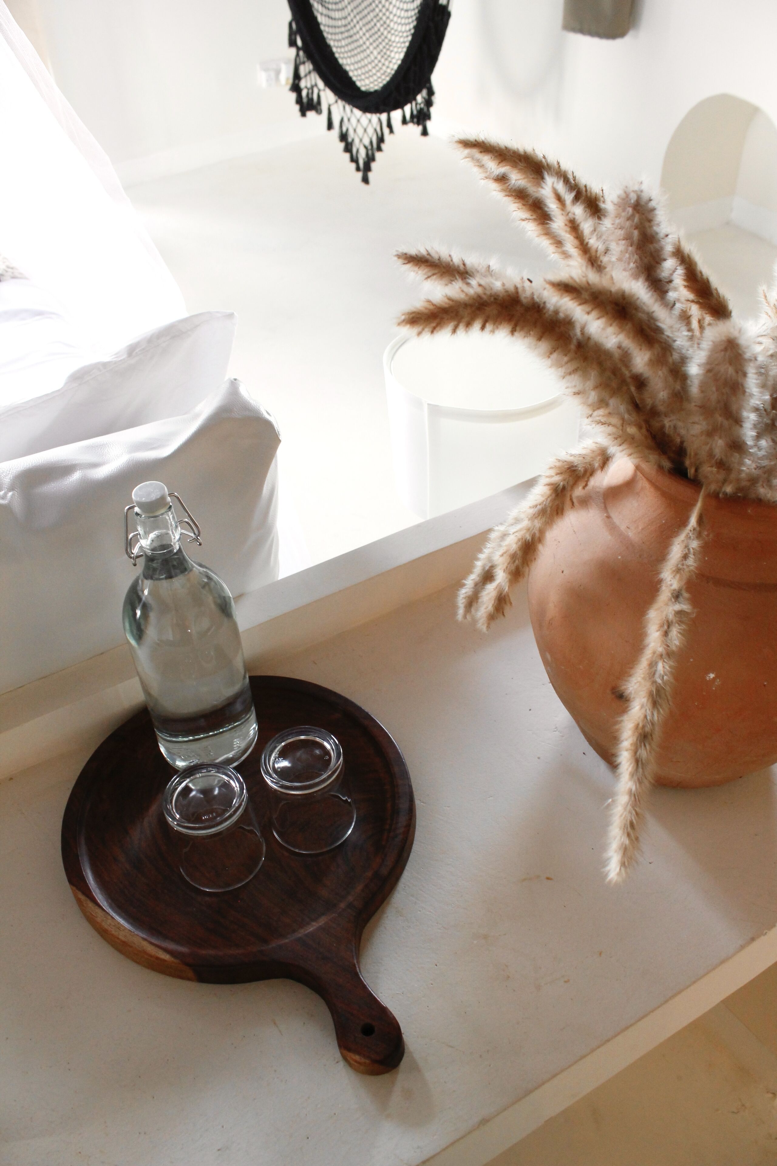 Een transparante glazen fles op een houten dienblad, met daaronder drie lege glazen, een terracotta pot met pluizweidegras, en een witte stoel op de achtergrond.