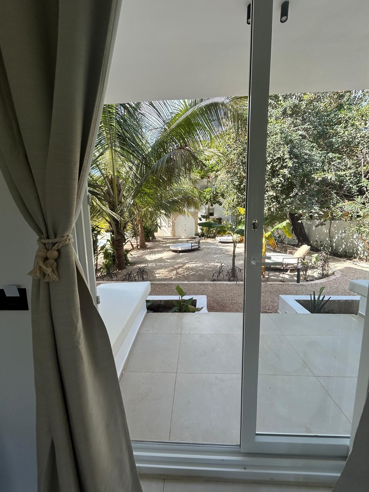 A view from a room with a sliding glass door leading to a patio area with a white outdoor seating arrangement, palm trees, and a small garden.