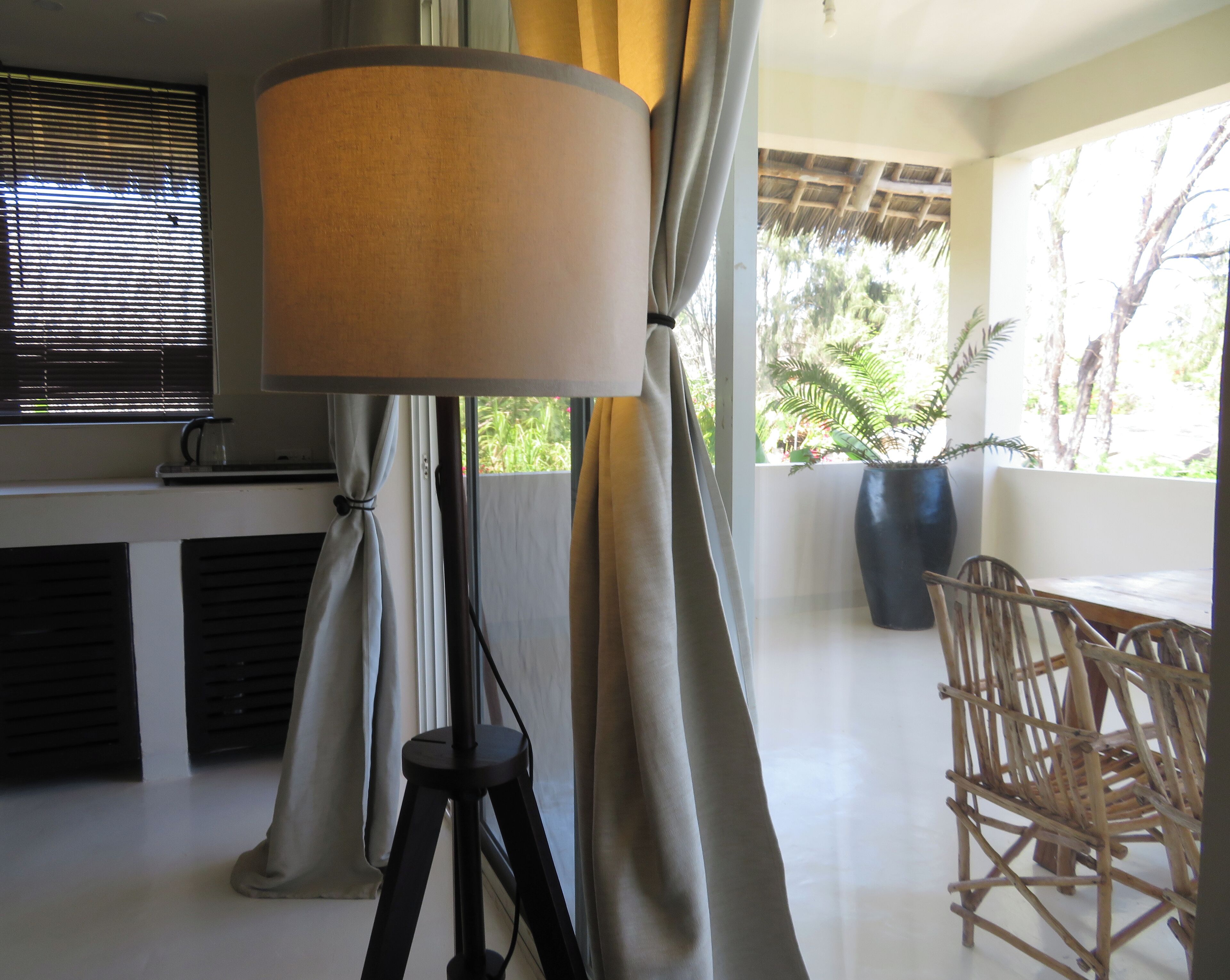 A room with a floor lamp, curtains, a table with chairs, a potted plant, and a view of greenery outside.