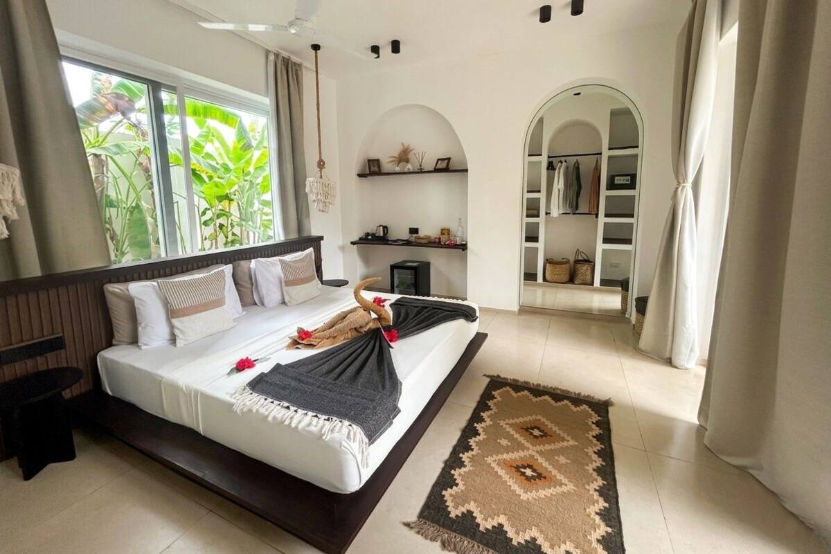 A bedroom with a bed, a bedside table, a nightstand, a window with curtains, a ceiling fan, a built-in shelf, and a wardrobe.