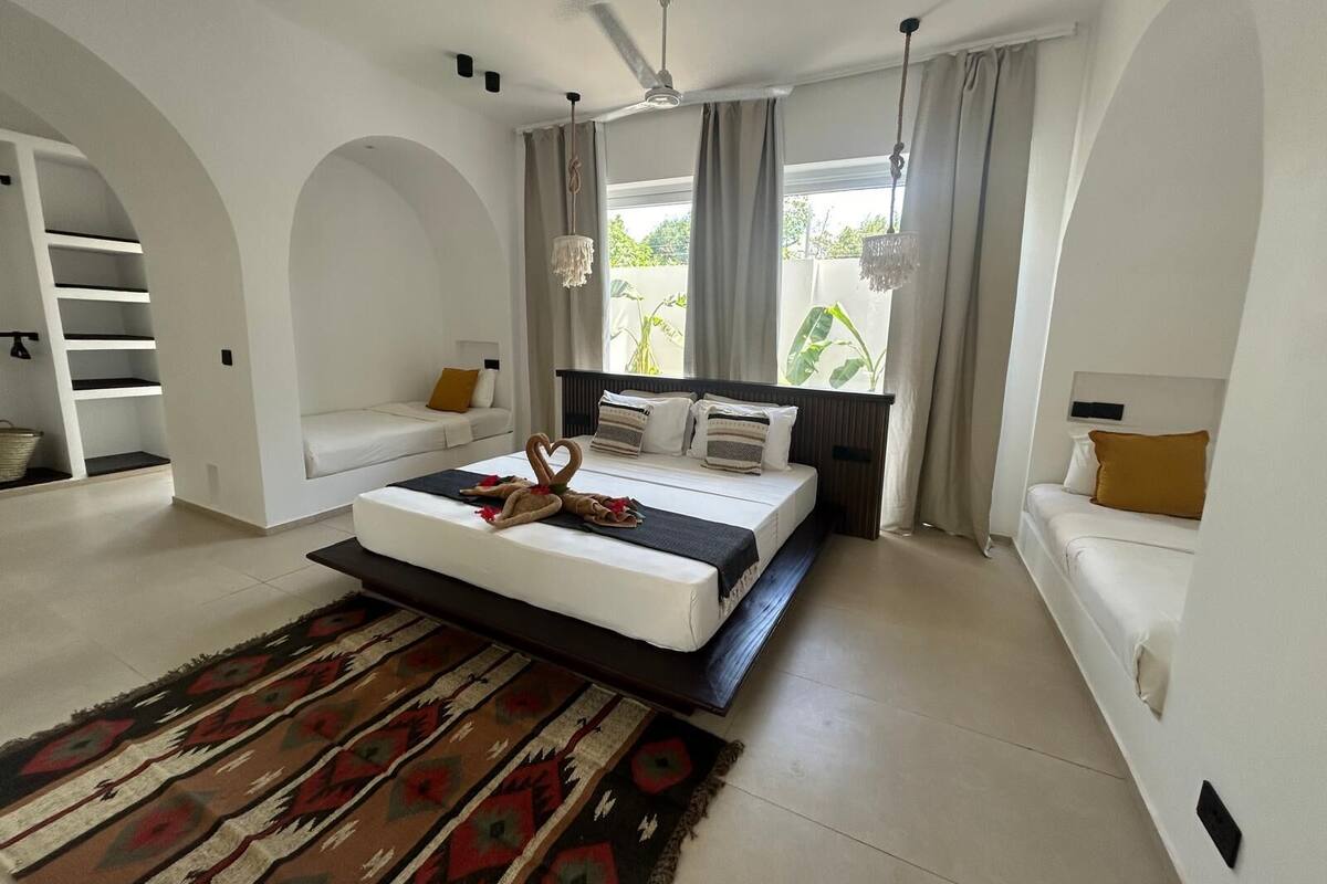 A modern bedroom with a large bed, a built-in bench, and a view of greenery outside.