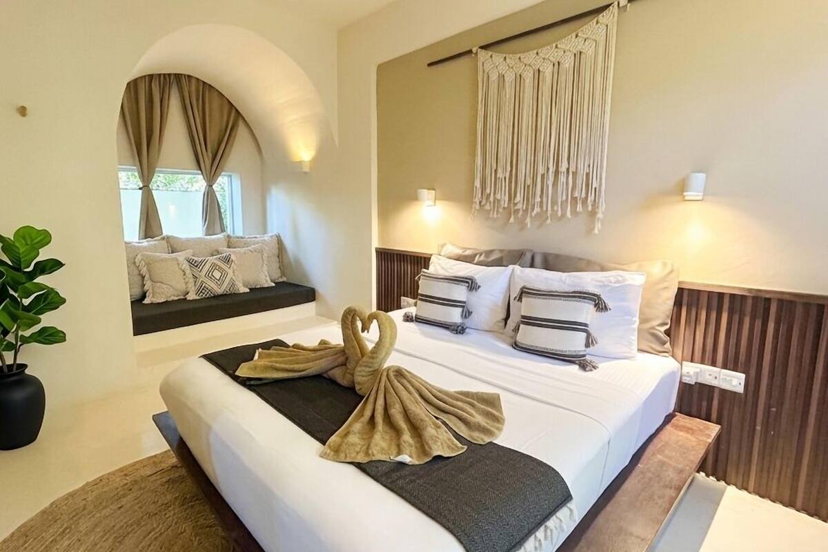A hotel room with a bed, a seating area, a potted plant, and a wall-mounted light fixture.