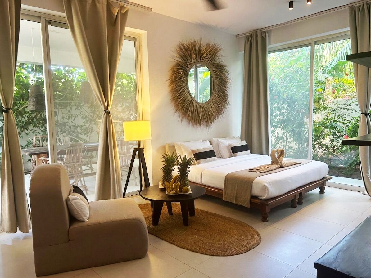 A bedroom with a bed, a sofa, a coffee table with fruit, a lamp, and a view of greenery outside.