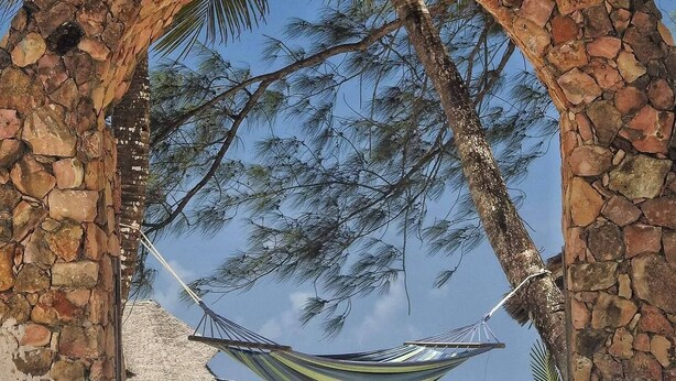 A stone archway with a hammock hanging inside, overlooking a beach with clear turquoise water and palm trees. - Kuwa Zanzibar (Kiwengwa)