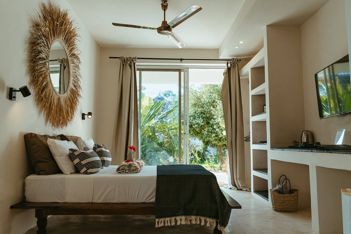 A bedroom with a bed, a ceiling fan, a large mirror, a TV, and a view of greenery through the window.