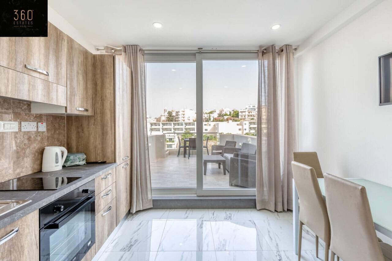 Une cuisine moderne avec des meubles en bois, un four encastré et une grande porte-fenêtre coulissante donnant sur un balcon aménagé avec du mobilier d’extérieur.