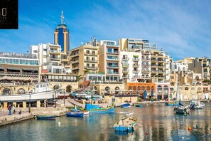 A coastal cityscape with a marina filled with boats, a prominent tower, and a variety of buildings including a Dolce Vita restaurant. - Beautiful, spacious APT with BBQ & private terrace (San Ġiljan)