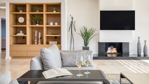 A modern living room with a sofa, a coffee table with an open book and two glasses of white wine, a large wooden bookshelf, a flat-screen TV, and a fireplace. - Jewel by AvantStay | Panoramic Ocean Views in La Jolla! (San Diego)