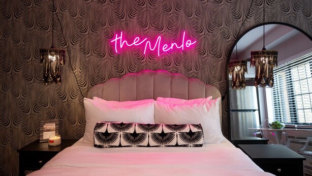 Un dormitorio moderno con un letrero de neón que dice 'theMenlo' sobre la cama, papel tapiz estampado y un espejo grande en la pared.