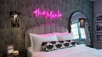 A bedroom with a tufted headboard, a neon sign reading "The Mento," two pendant lights, a bedside table with a candle, and a framed picture.