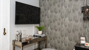 Un salón moderno con un televisor negro montado en la pared, un papel tapiz estampado, una consola con una planta, libros y una vela.