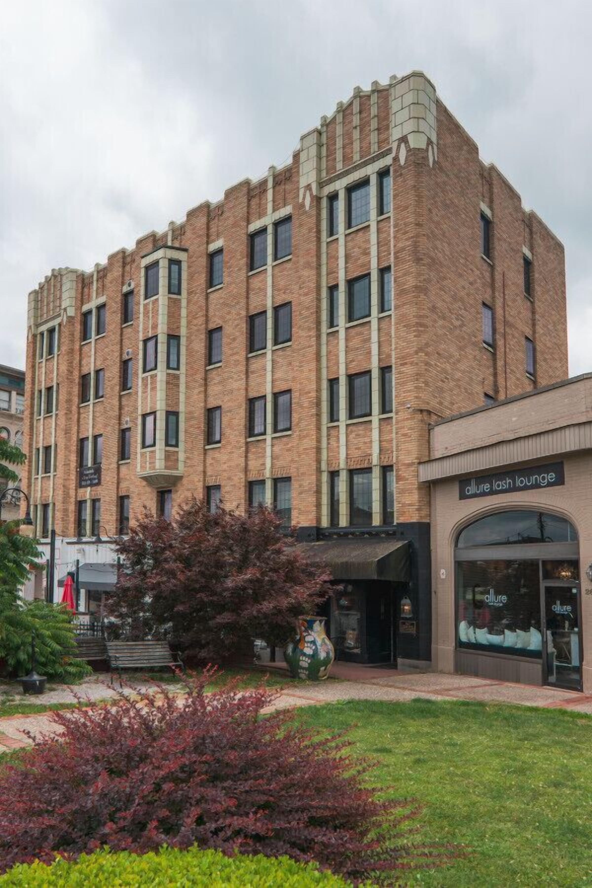 A multi-story building with a brick facade, a storefront labeled "Allure Lash Lounge," and a small garden area with shrubs and a bench.