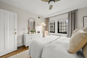 A bedroom with a bed, a dresser, a mirror, a chair, and two windows with curtains. - The Rookwood | Hyde Park (Cincinnati)