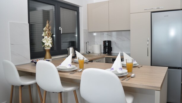 Una cocina moderna con una isla central, sillas blancas, un refrigerador y una ventana con persianas.