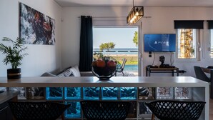 A modern living room with a large window, a dining area with a bowl of fruit, a flat-screen TV, and a potted plant. - Steps from Maleme beach, walking distance to shops, private pool! (Chania)