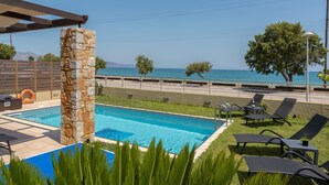A swimming pool with lounge chairs and a stone pillar, overlooking a body of water. - Steps from Maleme beach, walking distance to shops, private pool! (Chania)