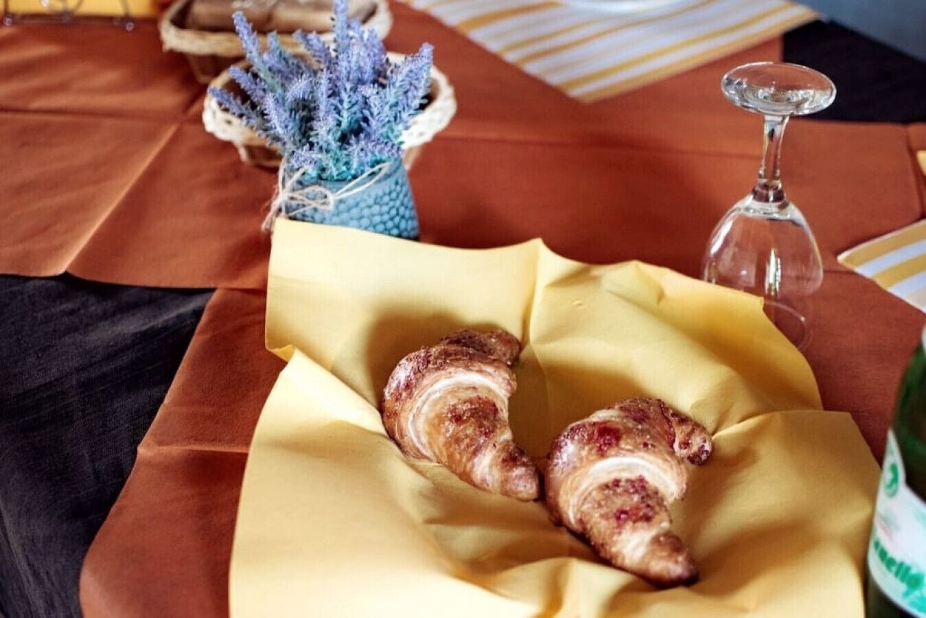 Dois croissants sobre um guardanapo amarelo, um copo de água e um vaso azul com lavanda.