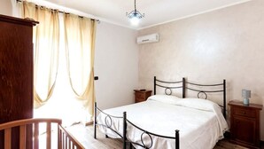 Un dormitorio con una cama, mesitas de noche de madera, una silla de madera, una ventana con cortinas y un equipo de aire acondicionado.